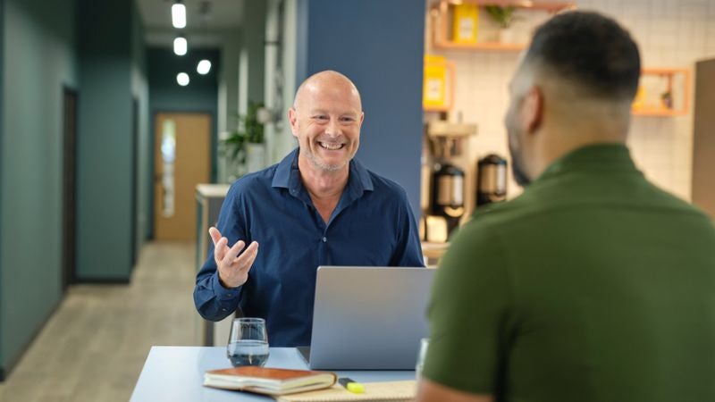 A man sat down with laptop smiling at another man across a table