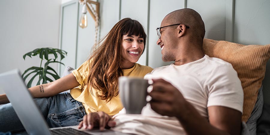 Woman and man with computer on lap smiling at each other sitting in bed.