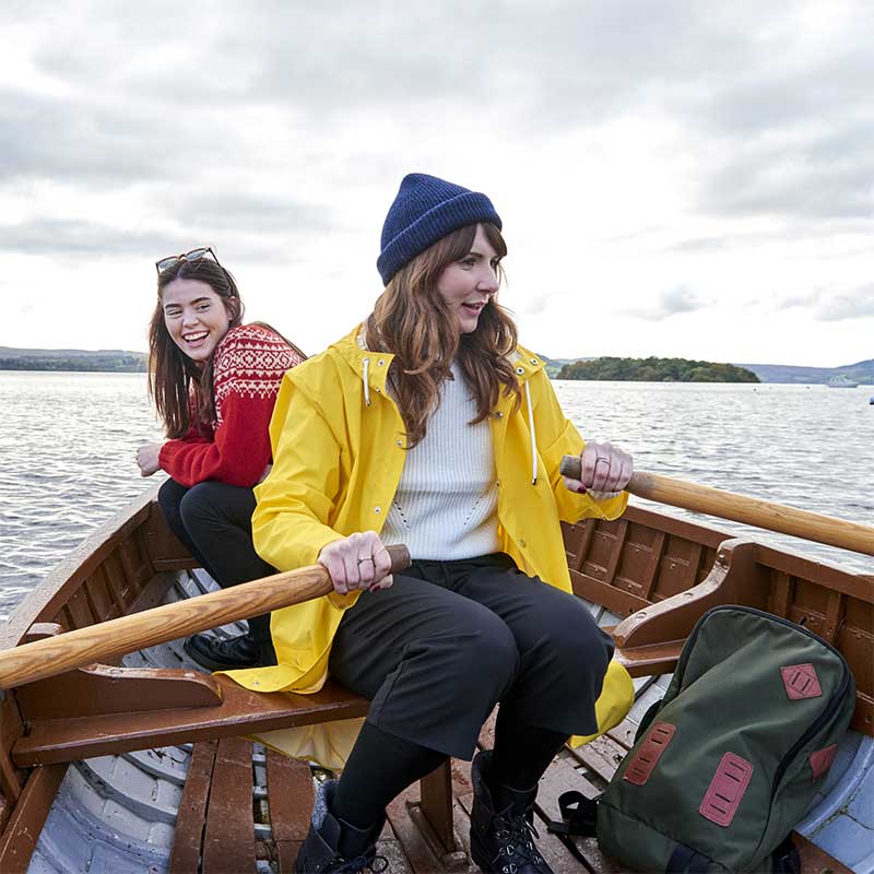 Two women in a row boat on a lake. One is wearing a yellow coat and blue hat, the other a red jumper.